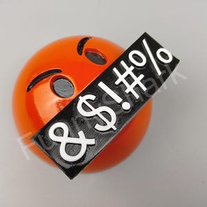 May include: An orange emoji with black eyes and a black rectangle covering its mouth. The rectangle has white text that includes an exclamation mark, a dollar sign, a hash, a percent sign, and an ampersand.
