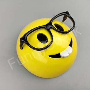 Puede incluir: Cara sonriente de cer&aacute;mica amarilla con gafas de montura negra y una amplia sonrisa.