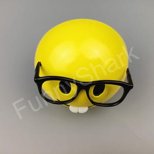 Puede incluir: Una cara sonriente amarilla con gafas de borde negro y una amplia sonrisa. La cara sonriente es un objeto en 3D.