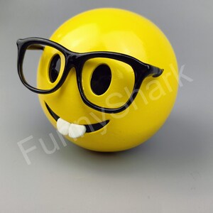 Puede incluir: Una cara sonriente amarilla con una sonrisa de dientes y gafas de montura negra.