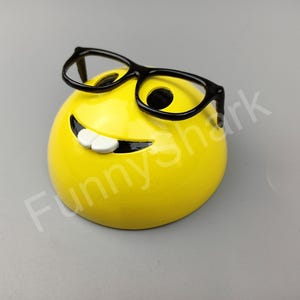 Puede incluir: Una cara sonriente amarilla con una amplia sonrisa y gafas de montura negra. La cara sonriente es una figurita de cer&aacute;mica.