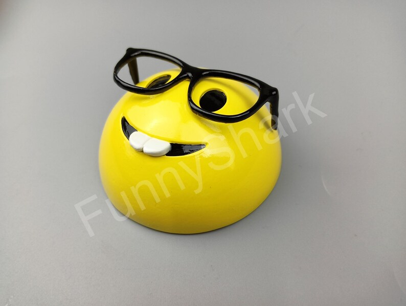 Puede incluir: Una cara sonriente amarilla con una amplia sonrisa y gafas de montura negra. La cara sonriente es una figurita de cer&aacute;mica.