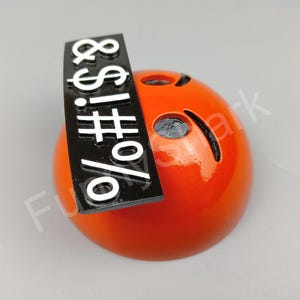 May include: A glossy orange sphere with cut-out eyes and a curved mouth. A black rectangular sign with white symbols and characters is attached to the top of the sphere.
