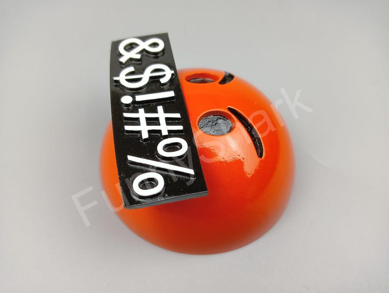 May include: A glossy orange sphere with cut-out eyes and a curved mouth. A black rectangular sign with white symbols and characters is attached to the top of the sphere.
