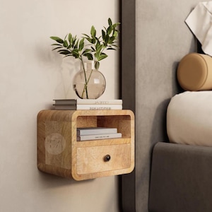 May include: A light wood floating bedside table with a drawer and a shelf. The bedside table is mounted to a white wall and has a small vase with green plants on top of it. The bedside table is next to a grey upholstered bed with white bedding.