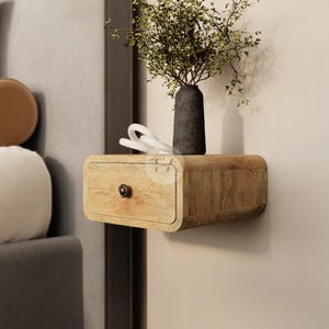Op de afbeelding: Een zwevend nachtkastje van hout met een enkele lade, aan de muur bevestigd. Een kleine, donkere vaas met groen staat erop. Een wit decoratief object ligt ernaast. Een hanglamp hangt erboven.