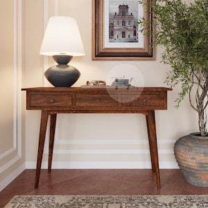 Può includere: Un tavolo console in legno scuro con tre cassetti e gambe affusolate. Una lampada bianca con un paralume nero è appoggiata sul tavolo, e una stampa incorniciata di un edificio è appesa al muro dietro di essa. Un ulivo in vaso con un vaso grigio si trova sul pavimento a destra del tavolo.