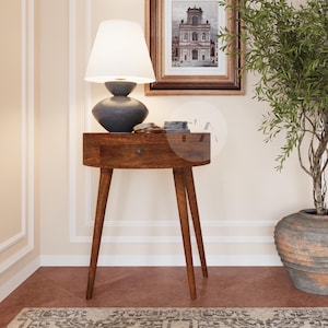 May include: A wooden nightstand with a lamp, artwork, and a potted plant. The nightstand has a drawer and tapered legs. The lamp has a white shade and a dark blue base. The artwork is framed in gold. A large potted plant is to the right.