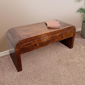 Può includere: Un tavolino in legno marrone scuro con due cassetti e pomelli in ottone. Il tavolo ha un design curvo ed è realizzato in legno massello. Due libri sono sopra il tavolo. Il tavolo è su un tappeto marrone.