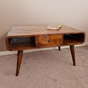 May include: A wooden coffee table with a drawer and two open shelves. The table has a dark brown finish and four tapered legs. A book is resting on the top of the table.