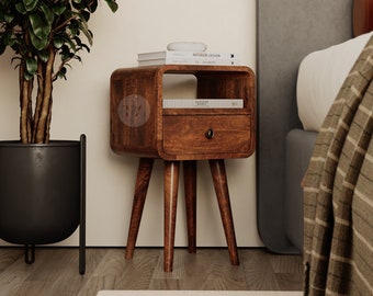 May include: A wooden bedside table with a drawer and a shelf. The table has four legs and is made of dark wood. The table is next to a bed with a gray headboard and a white blanket. There are books on the shelf and a plant in a black pot on the floor.