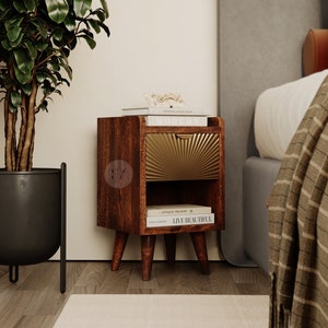 Op de afbeelding: Een houten nachtkastje met een gouden lade en een lager open vak. De tafel heeft vier poten en is versierd met boeken. De tafel staat in een slaapkamer met een bed en een plant.