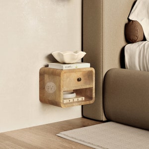 Può includere: Un comodino a parete in legno chiaro con un cassetto e uno scaffale. Il comodino è montato su una parete bianca accanto a un letto imbottito marrone.
