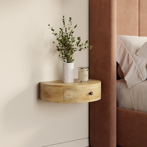 May include: A light wood floating shelf with a drawer and rounded edges. The shelf is mounted on a white wall and has a small potted plant and a candle on top. The shelf is next to a bed with a brown headboard and white bedding.