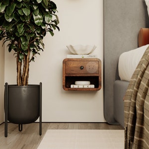 May include: A wall-mounted wooden bedside table with a single drawer and an open storage shelf below. The unit features a dark wood finish, a small black circular knob, and holds a white bowl and a book labelled ZOMA on the shelf.