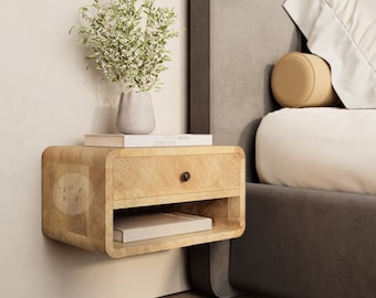 May include: A light wood floating nightstand with a drawer and a shelf. The nightstand is mounted on a white wall next to a gray upholstered bed with a white pillow and a tan throw pillow. A small vase with a green plant sits on top of the nightstand.