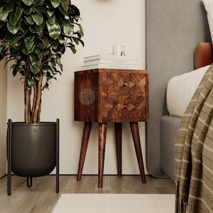 Op de afbeelding: Een donkerhouten nachtkastje met twee laden en vier poten. Het nachtkastje heeft een geometrisch patroon aan de voorkant en is bedekt met een stapel boeken en een glazen pot. Een groene, bladerrijke plant staat in een zwarte pot links van het nachtkastje.