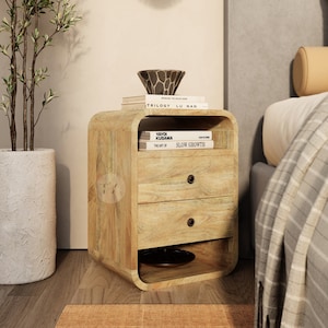 Peut inclure: Une table de chevet en bois avec deux tiroirs et une étagère. La table de chevet a un bord avant incurvé et est en bois brun clair. Il y a trois livres sur l'étagère supérieure, dont "Yayoi Kusama: The Art of Slow Growth".