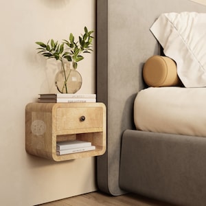 Peut inclure: Une table de chevet flottante en bois clair avec un tiroir et une étagère. La table de chevet est fixée à un mur blanc à côté d'un lit rembourré gris avec un oreiller blanc et un coussin de soutien-gorge beige. Un vase avec des plantes vertes feuillues est posé sur la table de chevet, et il y a trois livres empilés sur le vase.