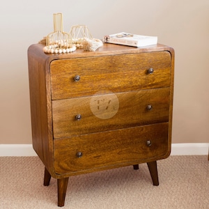 May include: A medium-brown wooden nightstand featuring three drawers with small, dark, circular knobs. The piece has rounded corners and is supported by four tapered legs. A book and decorative wire objects sit on the top surface of the unit.