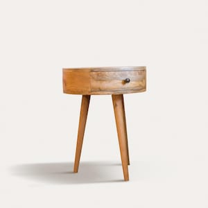 May include: A small, round wooden side table with a single drawer and three angled legs. The table is made of light-colored wood with a natural finish and a dark knob on the drawer. The design is mid-century modern.