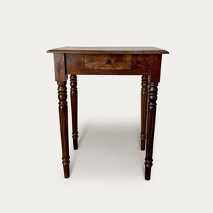 May include: A small, antique wooden table with a single drawer and turned legs. The table is a rich, dark brown color, and the drawer has a small, round knob. The legs are intricately carved, adding to the table's vintage charm.