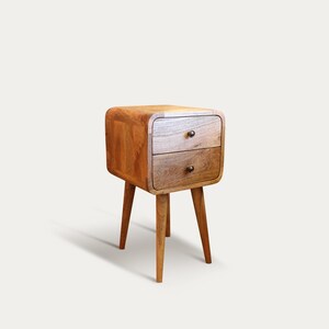 May include: A wooden nightstand with two drawers and rounded edges. The nightstand is supported by three angled legs. The wood has a warm, natural finish, and the drawer pulls are small, dark knobs.