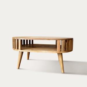 Könnte beinhalten: Ein hellbrauner Couchtisch aus Holz mit runder Oberseite und unterem Regal. Die Seiten weisen vertikale Lamellen auf, und der Tisch steht auf drei abgewinkelten Beinen. Minimalistisches und modernes Design.
