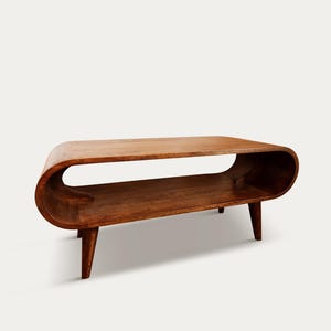 Peut inclure: Une table basse en bois de style moderne du milieu du siècle. Elle présente un design incurvé, une étagère inférieure et quatre pieds effilés. Le bois est de couleur marron chaud. La table mesure environ 122 cm de long, 46 cm de large et 41 cm de haut.