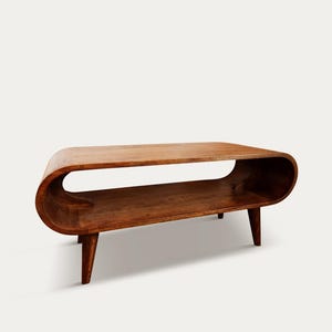 Peut inclure: Une table basse en bois de style moderne du milieu du siècle. Elle présente un design incurvé, une étagère inférieure et quatre pieds effilés. Le bois est de couleur marron chaud. La table mesure environ 122 cm de long, 46 cm de large et 41 cm de haut.