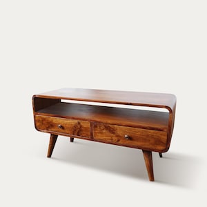 May include: A mid-century modern style wooden TV stand. The stand is a warm brown color with two drawers and a shelf. The stand has rounded edges and tapered legs. The drawers have small, round knobs.