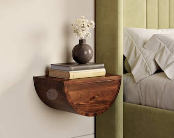 May include: A dark brown wooden half-circle nightstand with a drawer, mounted to a wall. The nightstand has a vase with white flowers and a stack of books on top. The nightstand is next to a green upholstered bed.