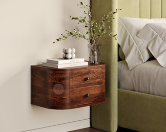 Pode incluir: Uma mesa de cabeceira flutuante de madeira castanho escura com duas gavetas e uma frente curva. A mesa de cabeceira está montada numa parede branca e tem um livro e objetos decorativos em cima.
