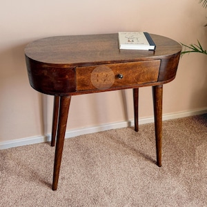 Può includere: Tavolino ovale in legno marrone scuro con un cassetto e quattro gambe affusolate. Un piccolo libro è appoggiato sopra. Il cassetto ha un pomello rotondo e scuro. Il tavolo è alto circa 76 cm.