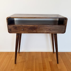 Può includere: Un tavolo consolle in legno marrone scuro caratterizzato da un piano rettangolare liscio, un ripiano a giorno e due cassetti con piccoli pomelli rotondi in metallo. Il mobile è sostenuto da quattro gambe affusolate e inclinate verso l'esterno.