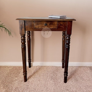 Peut inclure: Une table d'appoint en bois brun foncé avec un tiroir et des pieds tournés. La table a un carnet sur le dessus.