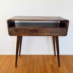 May include: A dark brown wooden console table featuring a smooth rectangular top, an open storage shelf, and two drawers with small round metal knobs. The piece is supported by four tapered, angled legs that extend outward from the base.