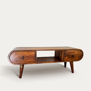Może przedstawiać: Drewniany stolik kawowy w stylu mid-century modern, z dwiema szufladami i półką. Stół ma ciepłe brązowe wykończenie, zaokrąglone krawędzie i zwężane nogi. Uchwyty szuflad to małe, ciemne gałki.