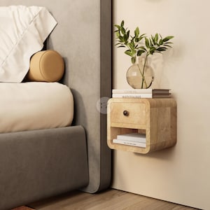 Op de afbeelding: Een zwevend nachtkastje van licht hout met een lade en een open plank, gemonteerd op een beige muur. Een glazen vaas met groene bladeren staat erop, samen met twee boeken. Een bed met een grijs hoofdbord is gedeeltelijk zichtbaar.
