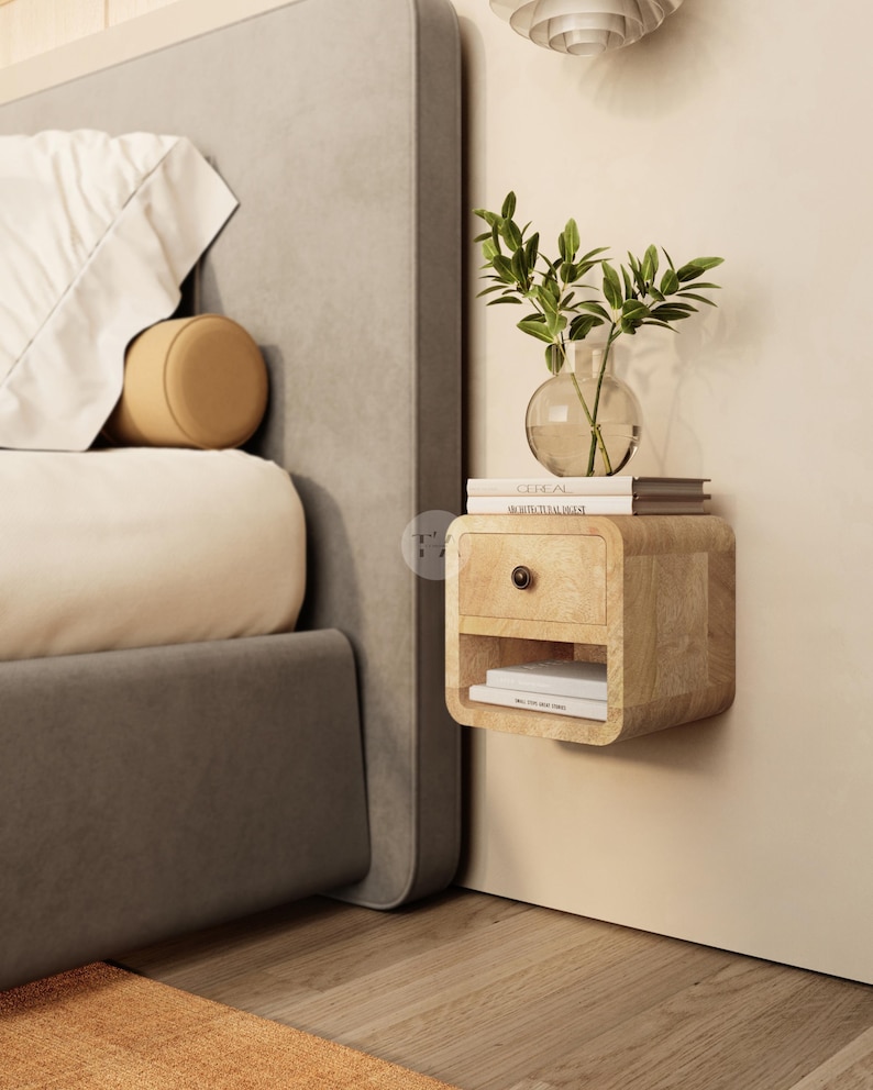 May include: A light wood floating nightstand with a drawer and open shelf, mounted on a beige wall. A glass vase with green leaves sits on top of the nightstand, along with two books. A bed with a gray headboard is partially visible.