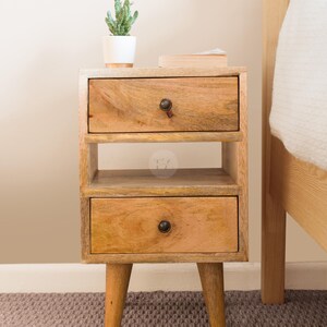 K&ouml;nnte beinhalten: Ein Nachttisch aus Holz mit zwei Schubladen und einem Regal. Der Tisch hat eine hellbraune Oberfl&auml;che und steht auf vier kurzen Beinen. Eine kleine Topfpflanze steht auf dem Tisch.