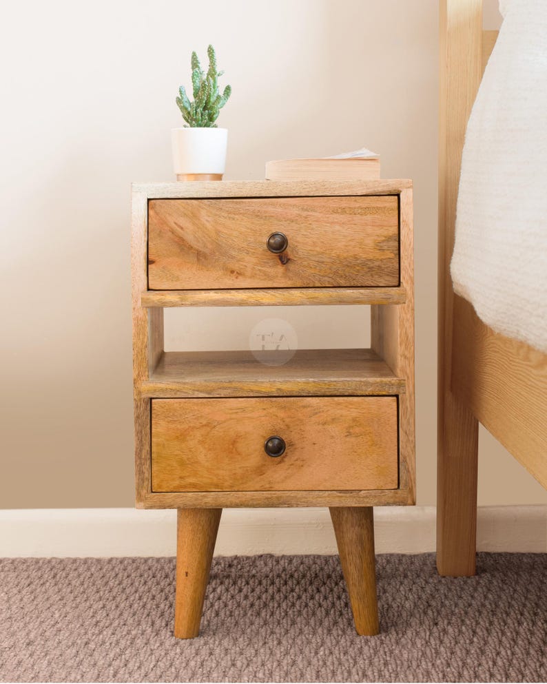 K&ouml;nnte beinhalten: Ein Nachttisch aus Holz mit zwei Schubladen und einem Regal. Der Tisch hat eine hellbraune Oberfl&auml;che und steht auf vier kurzen Beinen. Eine kleine Topfpflanze steht auf dem Tisch.
