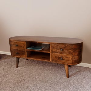 Può includere: Un mobile porta TV in legno marrone scuro con bordi arrotondati, dotato di quattro cassetti con piccoli pomelli scuri e due ripiani centrali aperti. Il mobile poggia su quattro gambe affusolate e presenta una finitura lucida con venature.