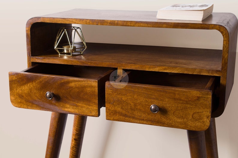 May include: A wooden side table with two drawers and a shelf. The table has a curved top and rounded edges. The drawers have metal knobs. The table is on a light background.
