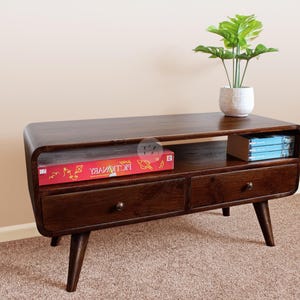May include: A dark brown wooden TV stand with two drawers and a shelf. The stand has rounded edges and tapered legs. A red game box and a stack of blue books are on the shelf. A potted plant sits on top.