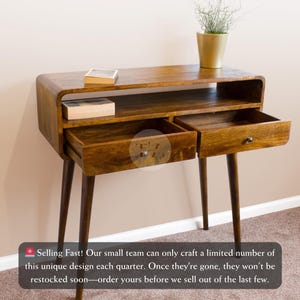 Puede incluir: Una consola de madera con dos cajones y un estante. La mesa tiene un acabado marrón cálido, patas cónicas y una parte superior curvada. Una pequeña planta en una maceta dorada está encima. Los cajones están abiertos.