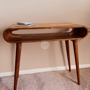 Puede incluir: Escritorio de madera de estilo moderno de mediados de siglo, con una parte superior curvada y un estante inferior. Tiene cuatro patas cónicas y un acabado de madera natural. Una pequeña pila de libros descansa sobre la superficie superior.