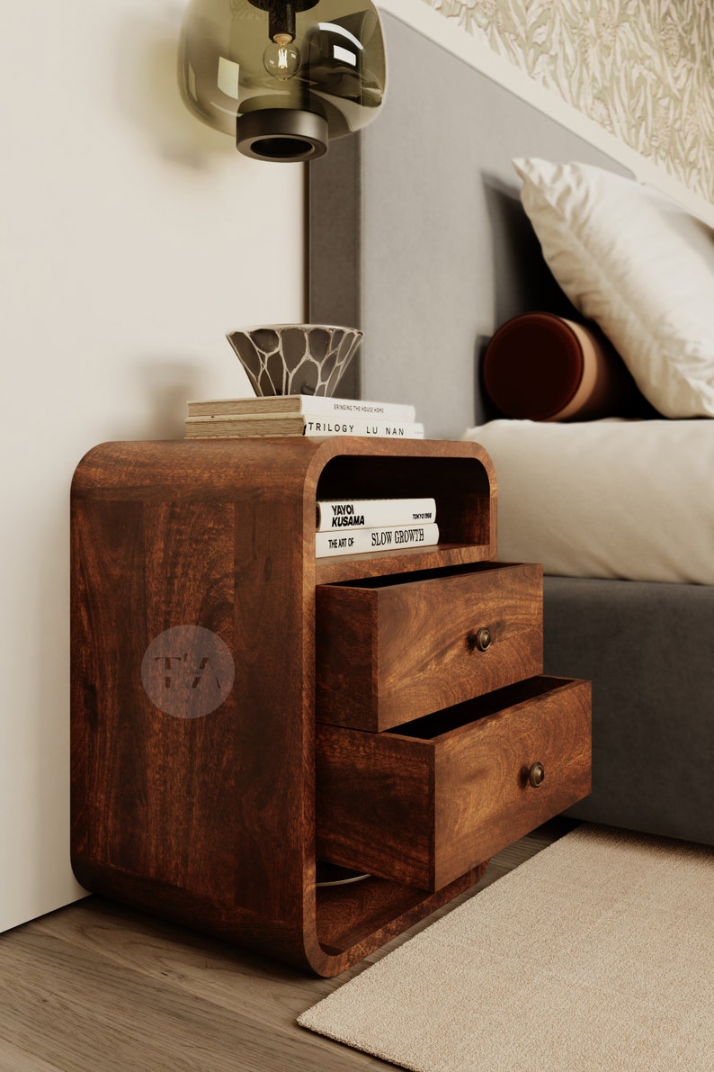 Pu&ograve; includere: Un comodino in legno marrone scuro con due cassetti e uno scaffale. Il comodino ha un design curvo e un'unica apertura sullo scaffale superiore. Il comodino &egrave; decorato con libri e una piccola ciotola.