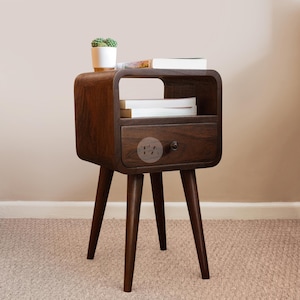 Può includere: Un comodino in legno marrone scuro con un cassetto e tre gambe angolate. Il comodino ha un piano arrotondato e un ripiano. Una piccola pianta in vaso e libri sono sopra. Il comodino è alto circa 61 cm.