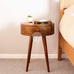 May include: A wooden bedside table with a single drawer and four legs. The table is brown and has a rounded top. A small potted plant and a book are on top of the table.